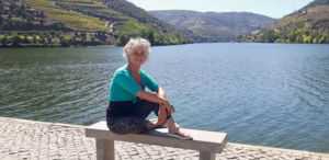 Imago-stylist Herma zit ontspannen aan het water met bergen op de achtergrond, moment van rust en reflectie.