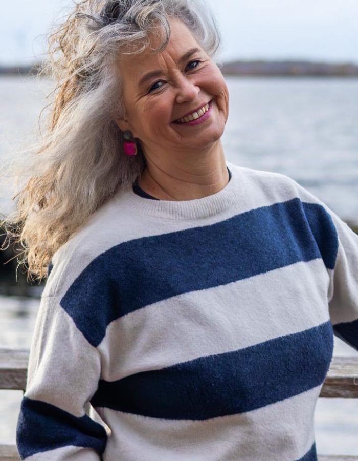 Herma staat buiten aan het water, draagt een lichtgekleurde trui met donkere strepen en oorbellen.