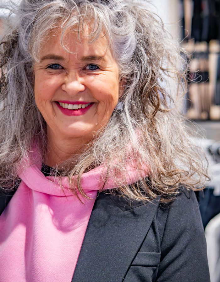 Portret van Herma, glimlachend, met grijs krullend haar, roze top en donker jasje, in een winkelomgeving.